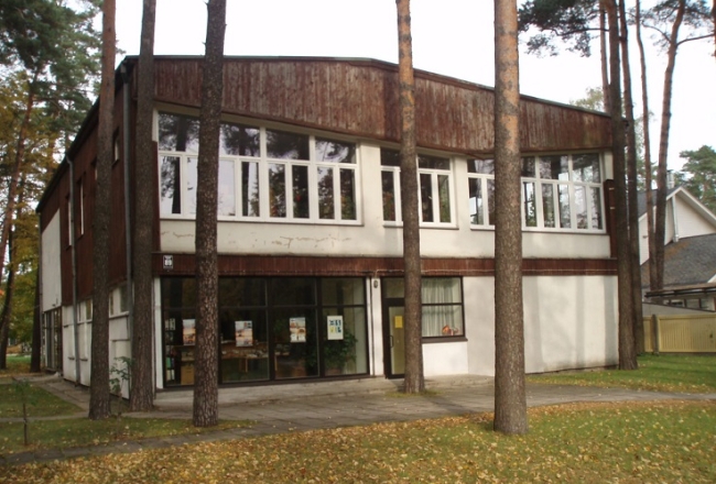 Divstāvīga plata ēka starp priedēm. Ēkas kreisajā pusē stiklotas durvis.