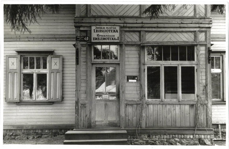 Bibliotēkas galvenās durvis, katrā pusē logs. Virs durvīm uzraksts latviešu un krievu valodā "Jūrmalas pilsētas 1. bibliotēka".