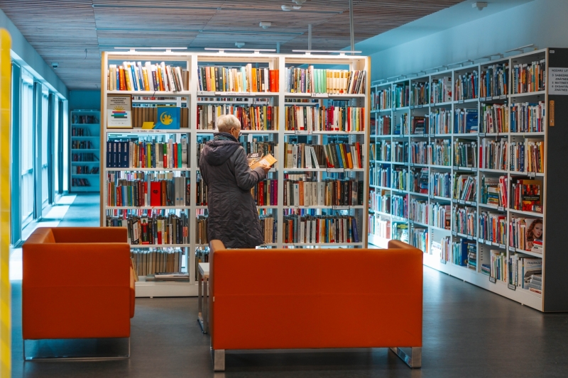 Sieviete pēta grāmatas pie plaukta Jūrmalas Centrālajā bibliotēkā, priekšplānā oranži dīvāni.