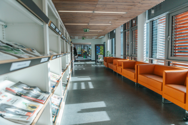 Bibliotēkas žurnālu un laikrakstu plaukti un oranži krēsli lasīšanai