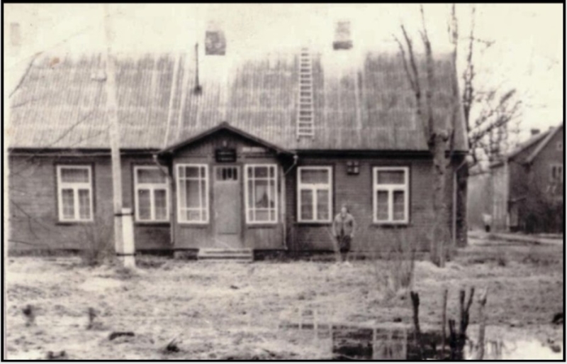 Vēsturiska melnbalta fotogrāfija ar koka ēku un personu tās priekšā.