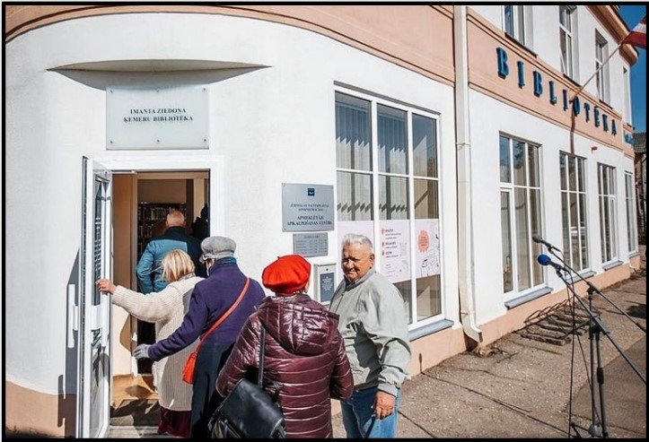 Pieci cilvēki dodas iekšā bibliotēkā. Bibliotēka ir divstāvīga ēka ar lieliem logiem un durvīm ēkas stūrī.