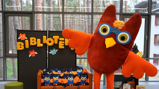 Liels brūni oranžs pūces maskots ar brillēm stāv bibliotēkā blakus stendam ar uzrakstu "BIBLIOTĒKA" un dīvānam, kas pilns ar pūču dizaina mugursomām. Foto Sanita Kitajeva