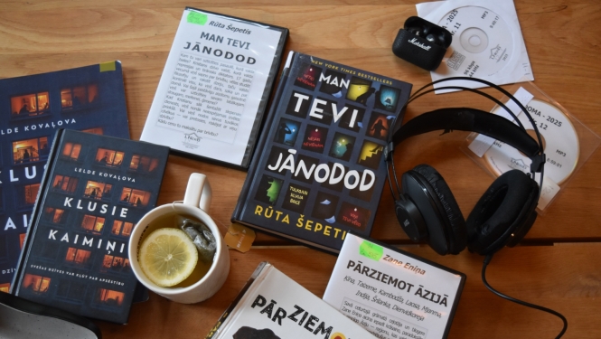 Skats no augšas uz koka galda izkārtotām grāmatām latviešu valodā, audiogrāmatu diskiem, austiņām, brillēm un tasi tējas ar citronu