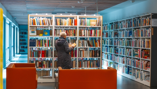 Sieviete pēta grāmatas pie plaukta Jūrmalas Centrālajā bibliotēkā, priekšplānā oranži dīvāni.