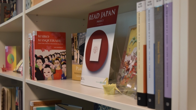 Grāmatu plaukts ar grāmatām par Japānas kultūru, izstādes 'Read Japan Project' plakātu un nelielu origami dzērvīti.
