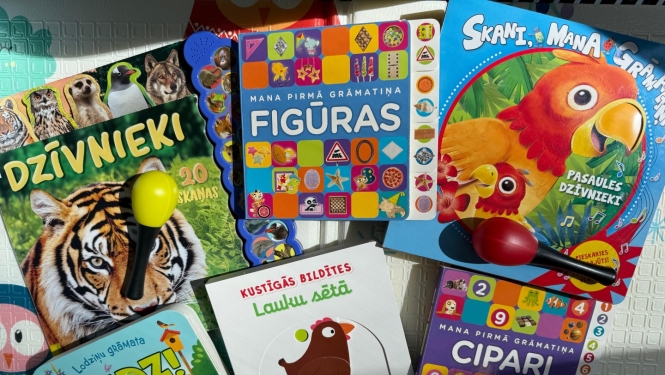 Krāsains bērnu grāmatu un rotaļlietu izkārtojums. Attēlā redzamas sešas dažādas bērnu izglītojošās un interaktīvās grāmatas latviešu valodā ("Dzīvnieki", "Mana pirmā grāmatiņa. Figūras", "Skani, mana grāmatiņa! Pasaules dzīvnieki", "Kustīgās bildītes. Lauku sētā", "Lodziņu grāmata. Audz!", "Mana pirmā grāmatiņa. Cipari") un divas mazas marakas (mūzikas instrumenti) dzeltenā un sarkanā krāsā.