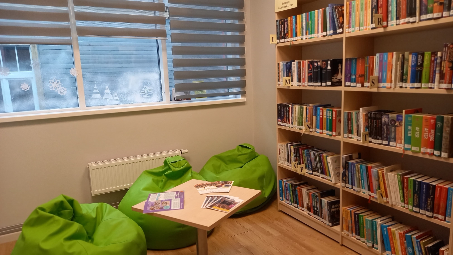 Mājīgs bibliotēkas lasīšanas stūrītis ar zaļiem sēžammaisiem, galdiņu un grāmatu plauktu.