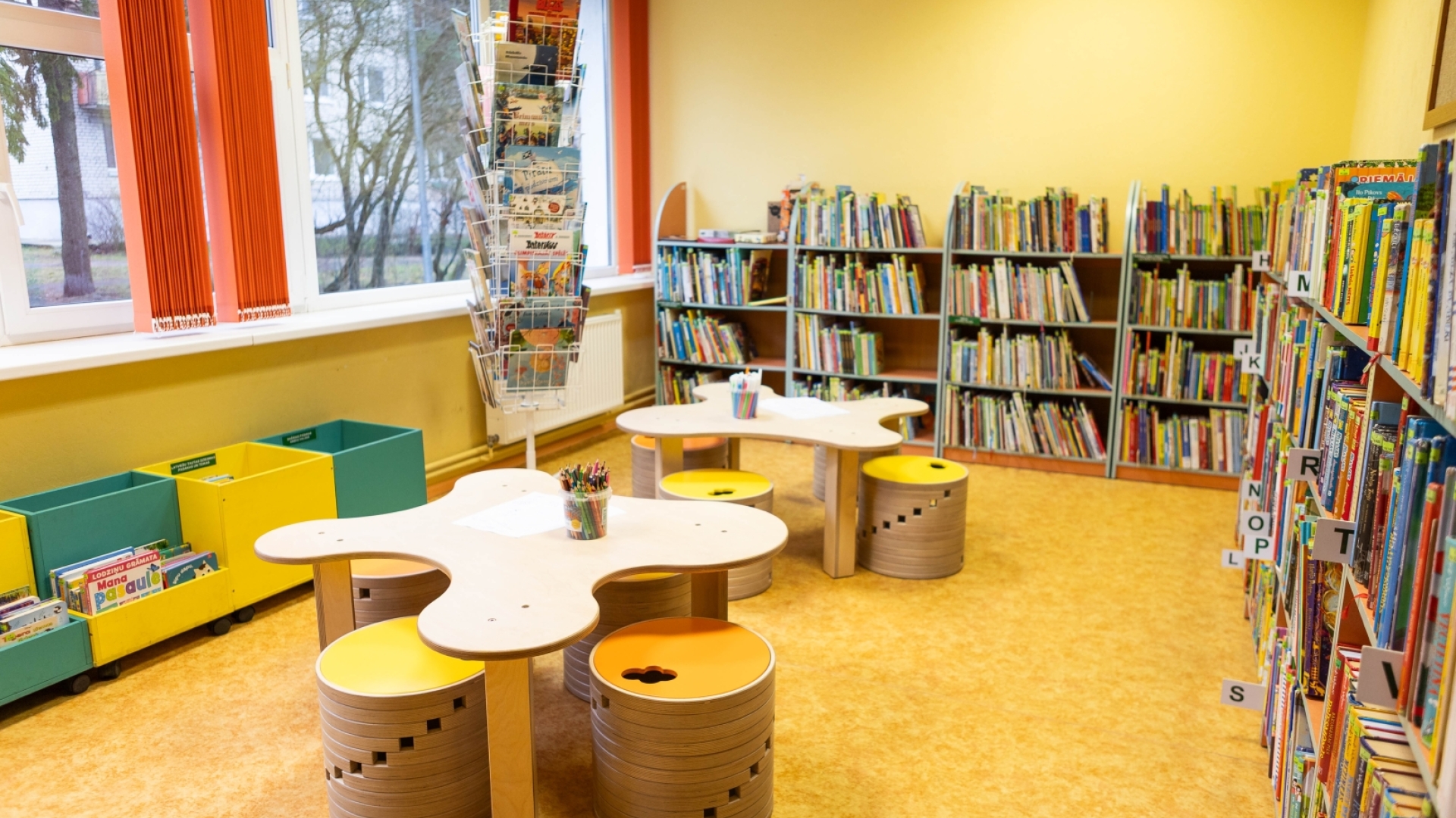 Radoša bērnu telpa bibliotēkā ar ābola formas galdiņiem, apaļiem krēsliem un daudziem plauktiem ar bērnu grāmatām