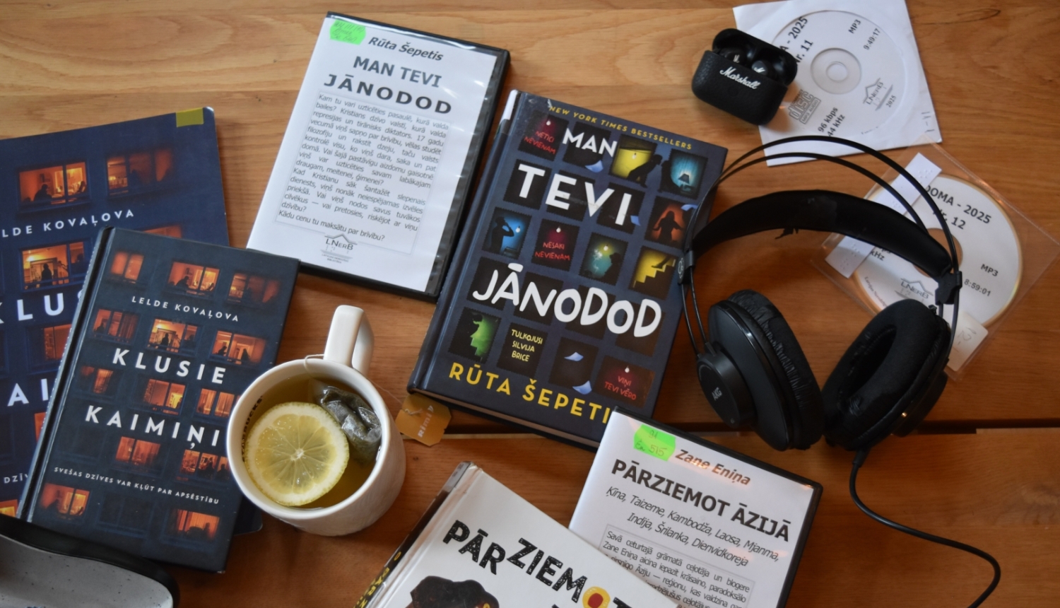 Skats no augšas uz koka galda izkārtotām grāmatām latviešu valodā, audiogrāmatu diskiem, austiņām, brillēm un tasi tējas ar citronu
