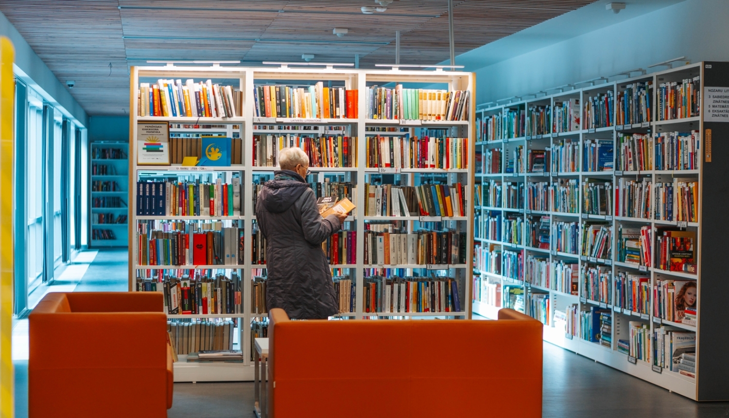 Sieviete pēta grāmatas pie plaukta Jūrmalas Centrālajā bibliotēkā, priekšplānā oranži dīvāni.