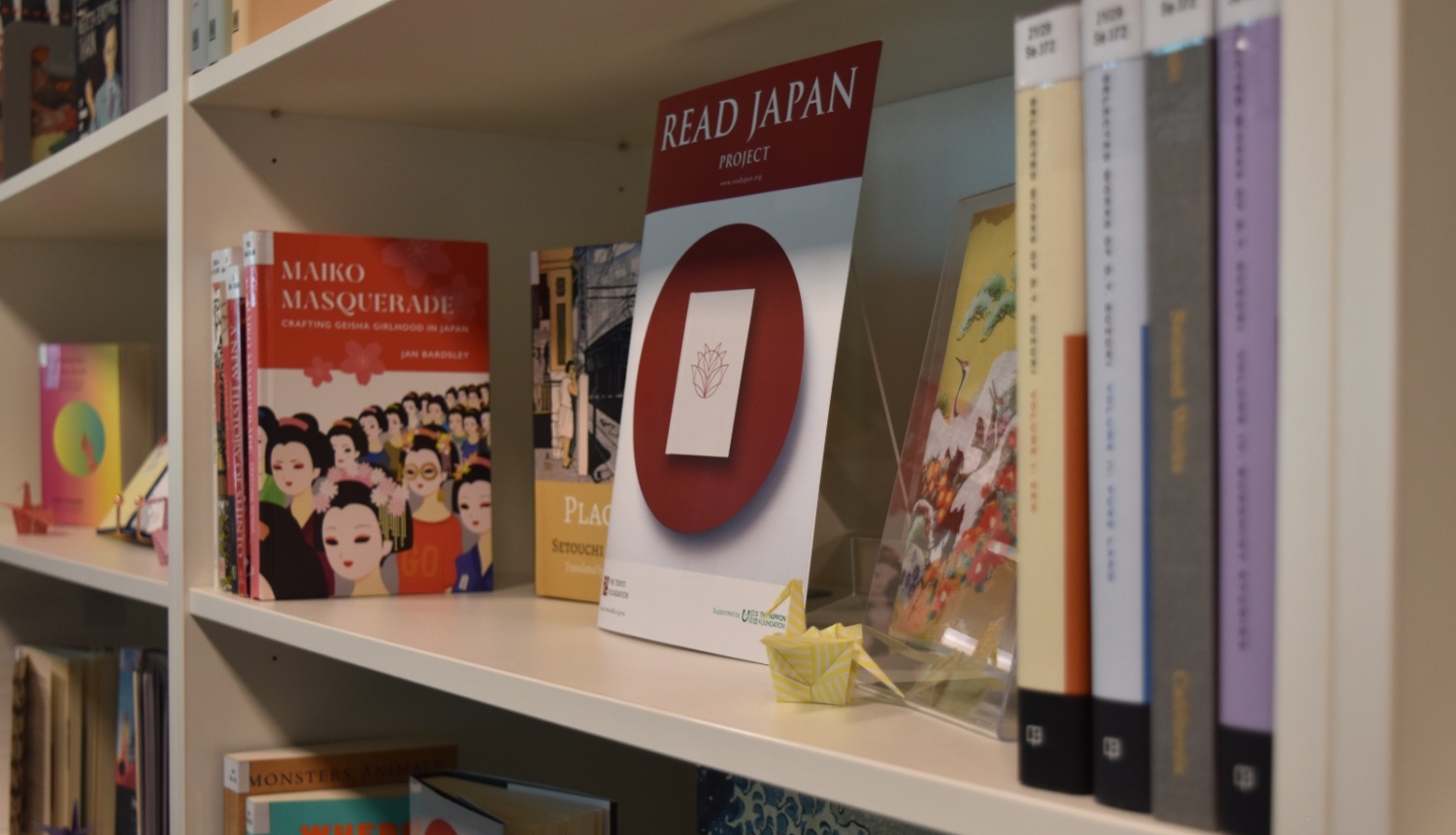 Grāmatu plaukts ar grāmatām par Japānas kultūru, izstādes 'Read Japan Project' plakātu un nelielu origami dzērvīti.